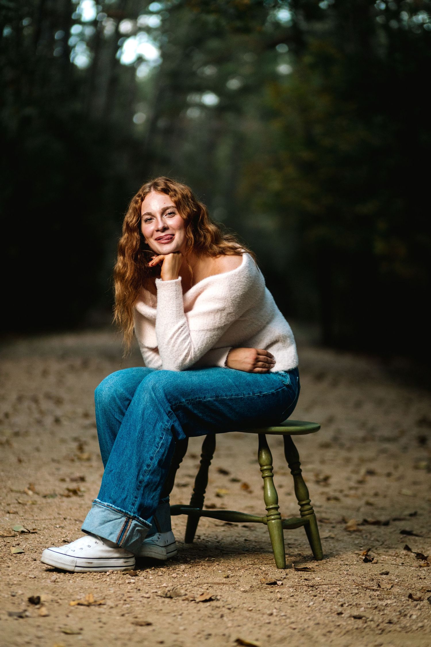 Person in white sweater and blue jeans sits casually on wooden stool against blurred forest background.