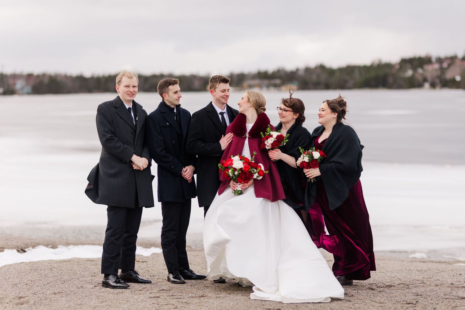 Wedding party in burgundy and black attire poses together on a snowy beach during winter celebration.