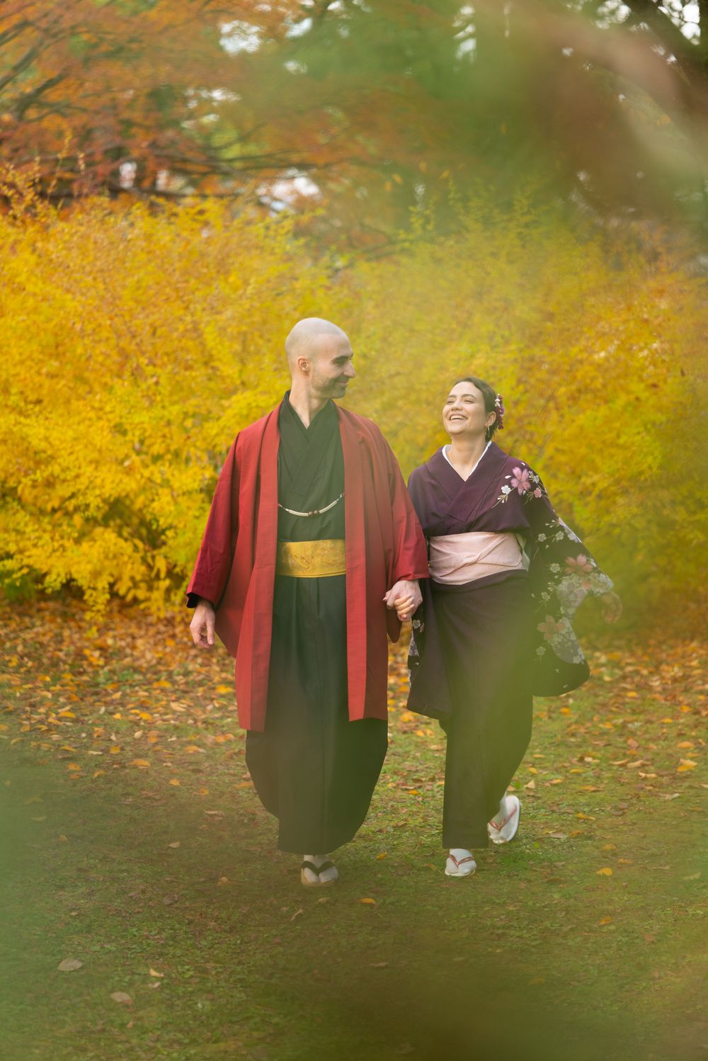 Couple in colorful kimono walking hand-in-hand through a vibrant autumn park with golden leaves.