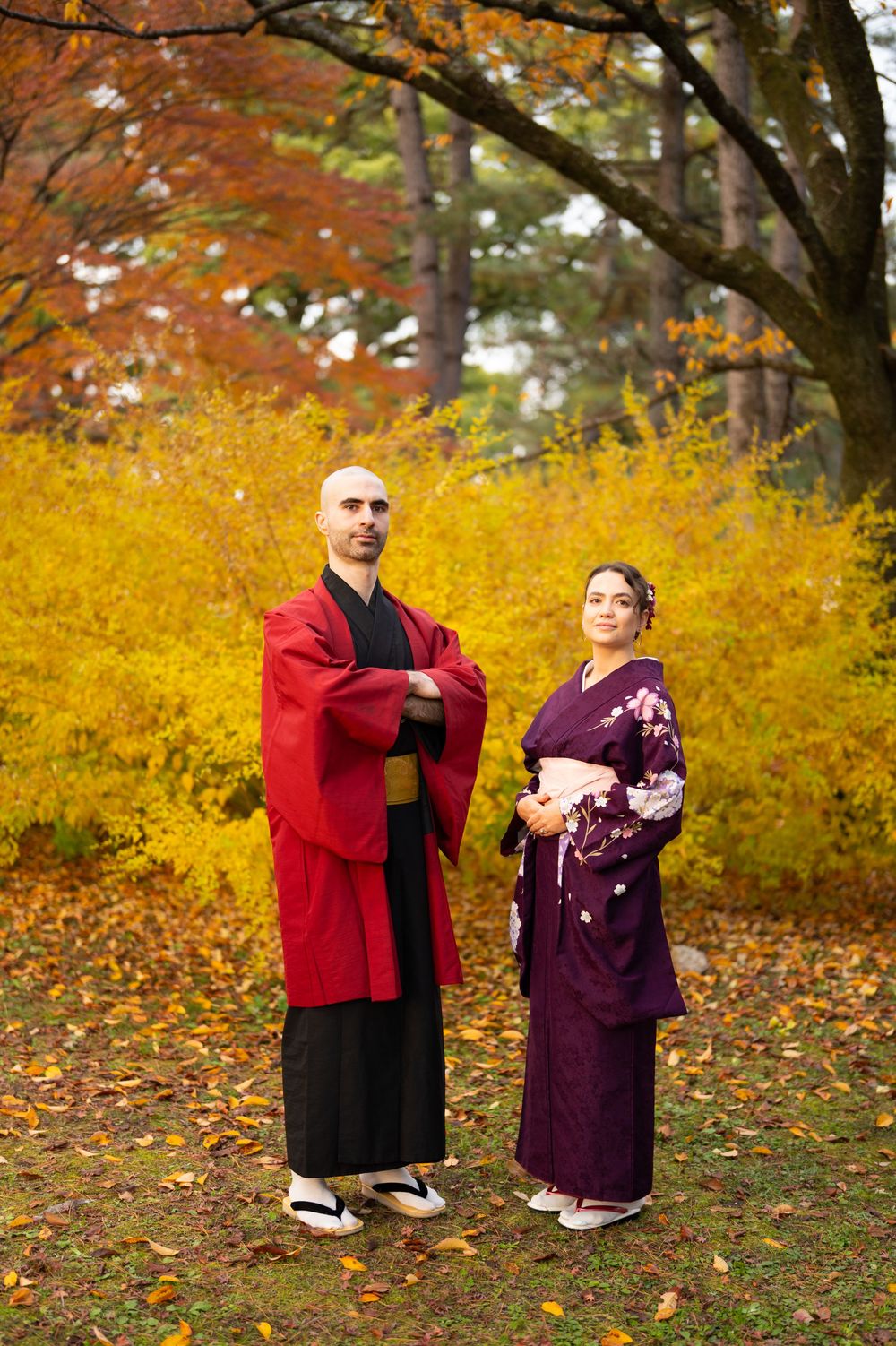 Man in red haori and woman in purple kimono standing together amid stunning autumn foliage.