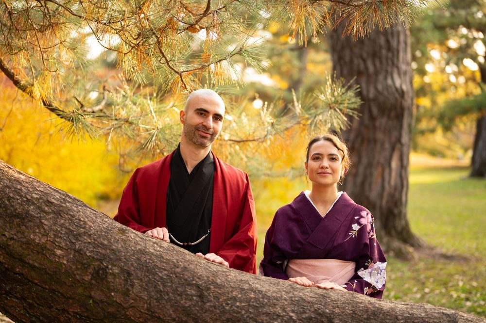 Couple in traditional Japanese kimono posing together beside a tree trunk in autumn park.