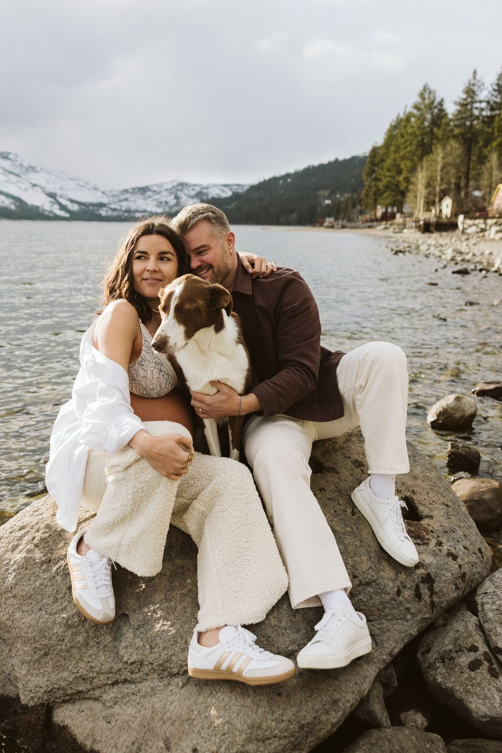 Couple in white outfits sitting with their dog on rocky shoreline with snow-capped mountains and lake in background.