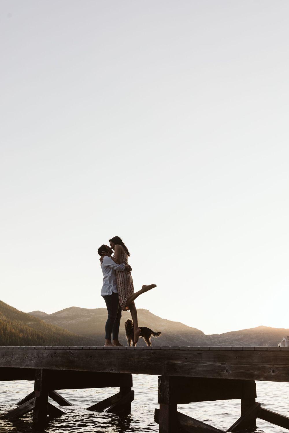 Silhouetted couple embracing on dock with dog against mountain landscape at dusk.