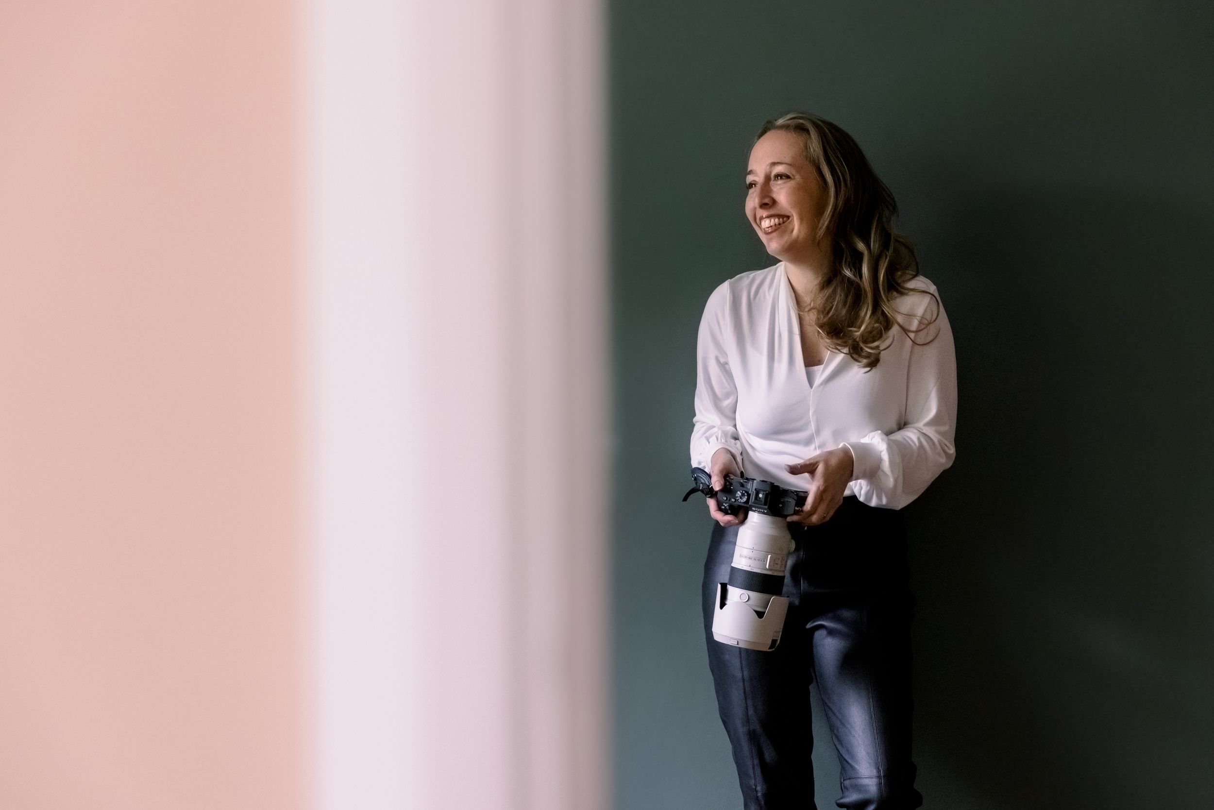 Person in white blouse holding camera and laughing against pink and green wall.