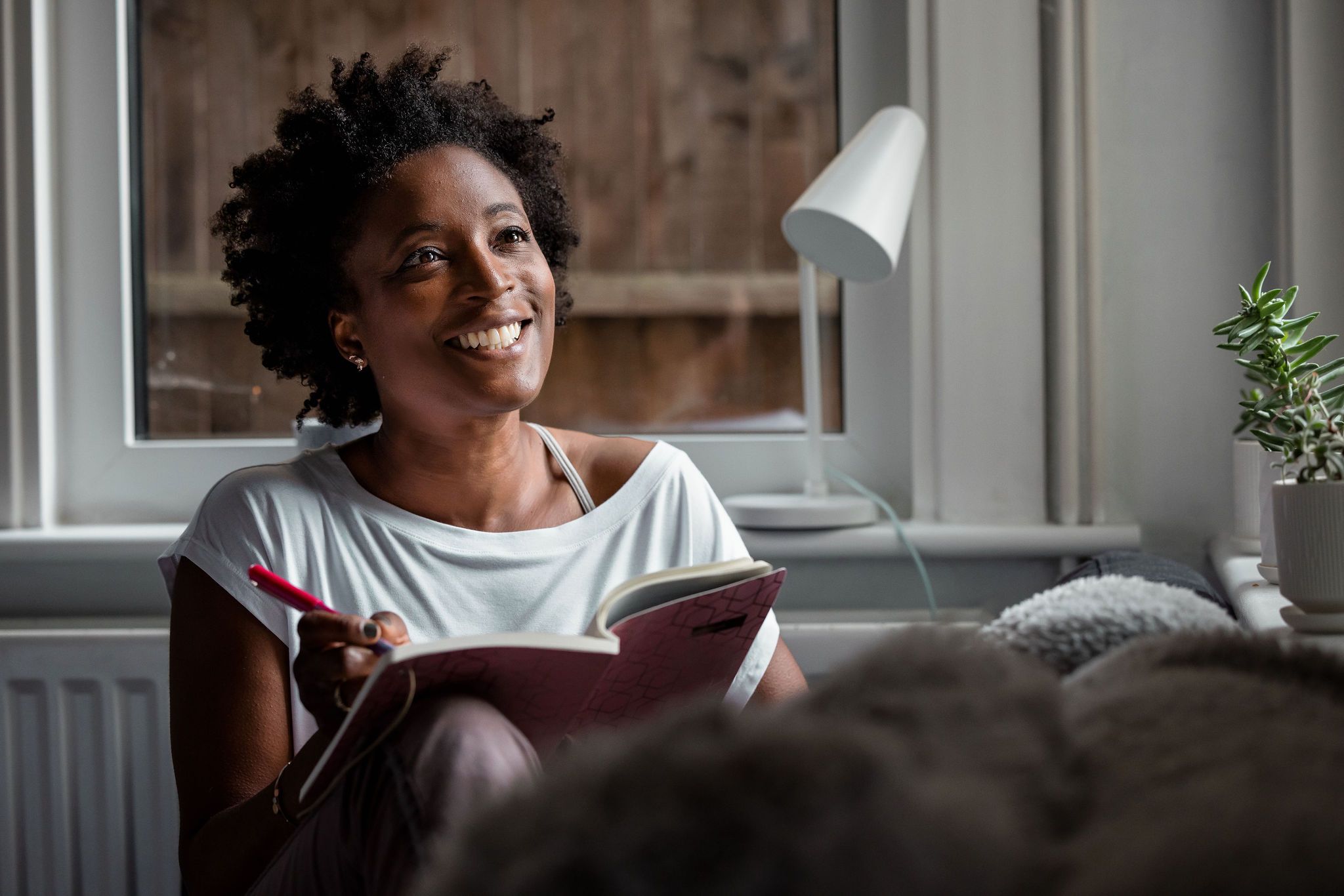 Person writing in notebook while relaxing on couch near window with desk lamp.