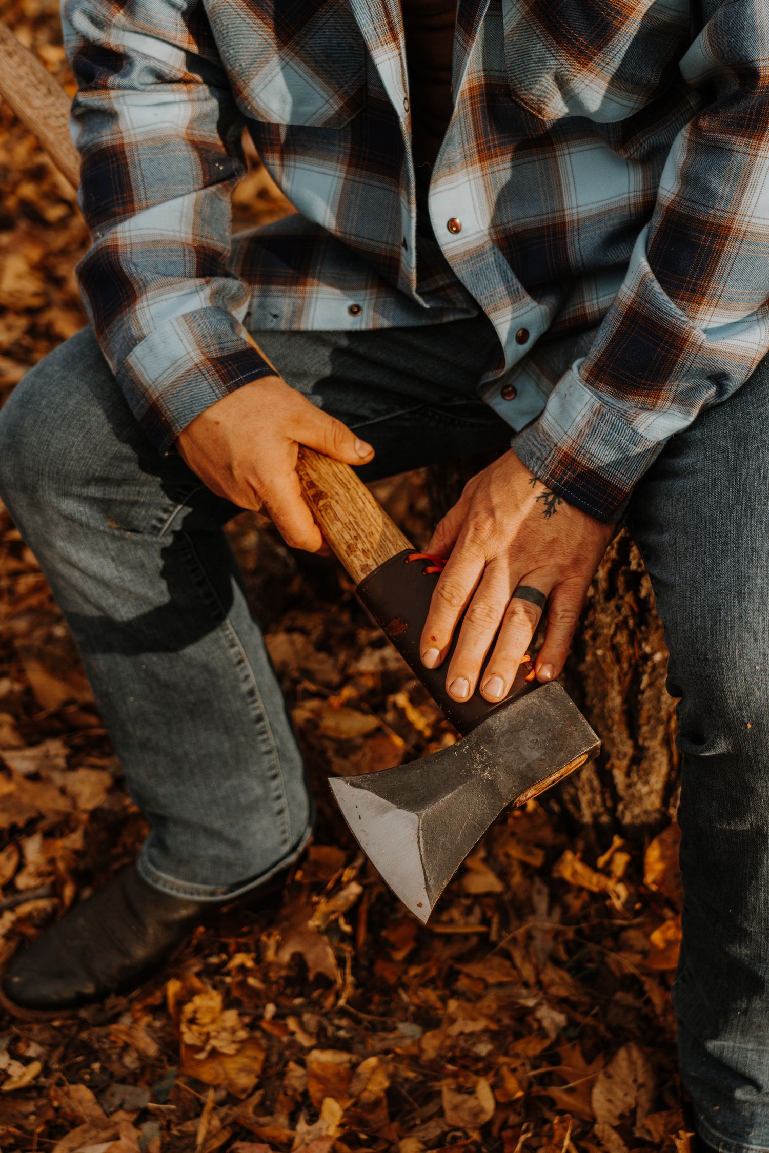 Hands holding an axe while sitting on a log surrounded by fallen autumn leaves.
