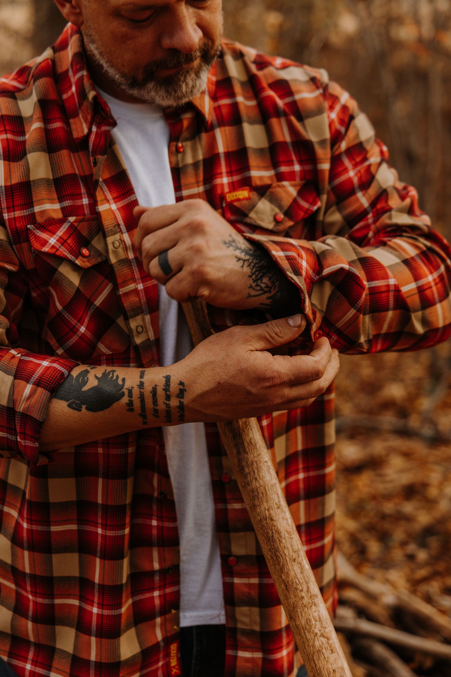Person wearing red plaid shirt carving wood with a knife in an autumn forest setting.