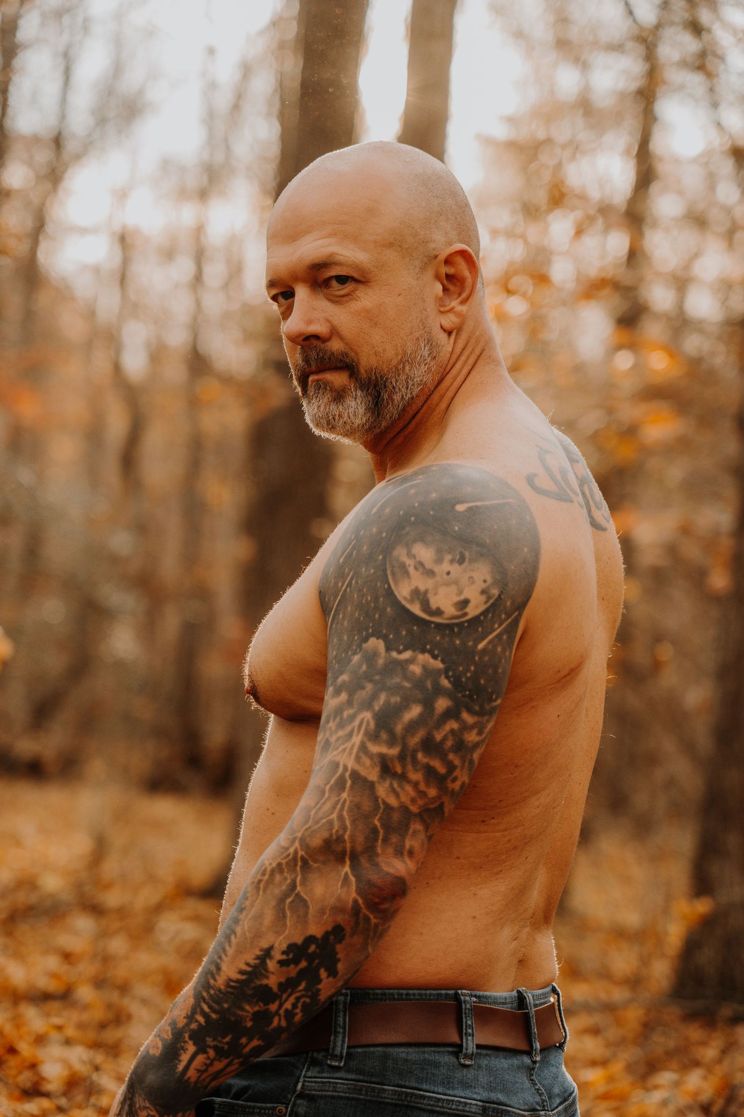 Shirtless person with arm tattoos stands in autumn forest during golden hour sunset.