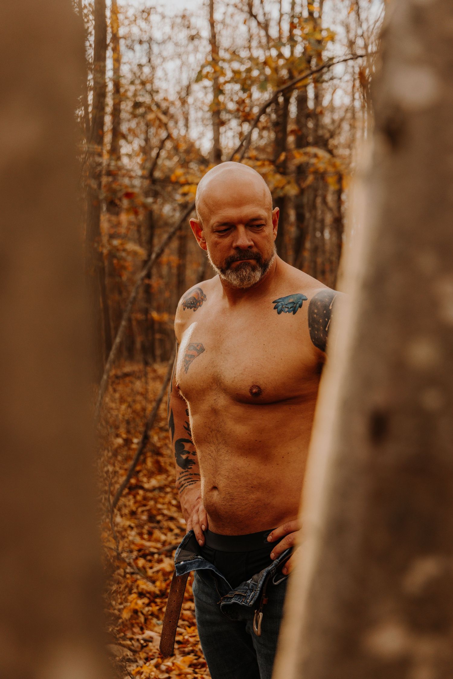 Shirtless person with tattoos stands among autumn trees in a moody forest setting during golden hour.