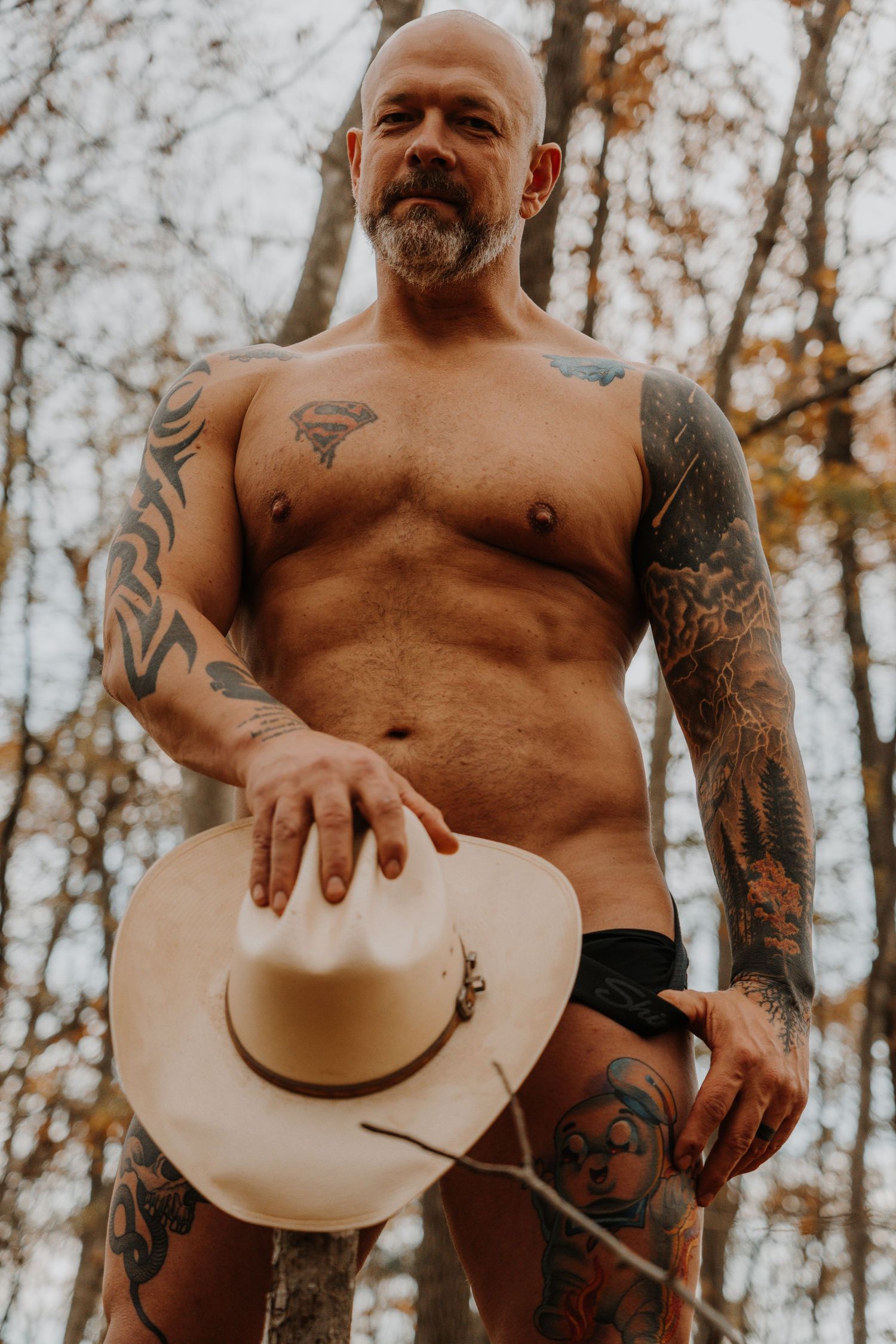 Artistic nude photograph of a tattooed person holding a cowboy hat in an autumn forest setting.