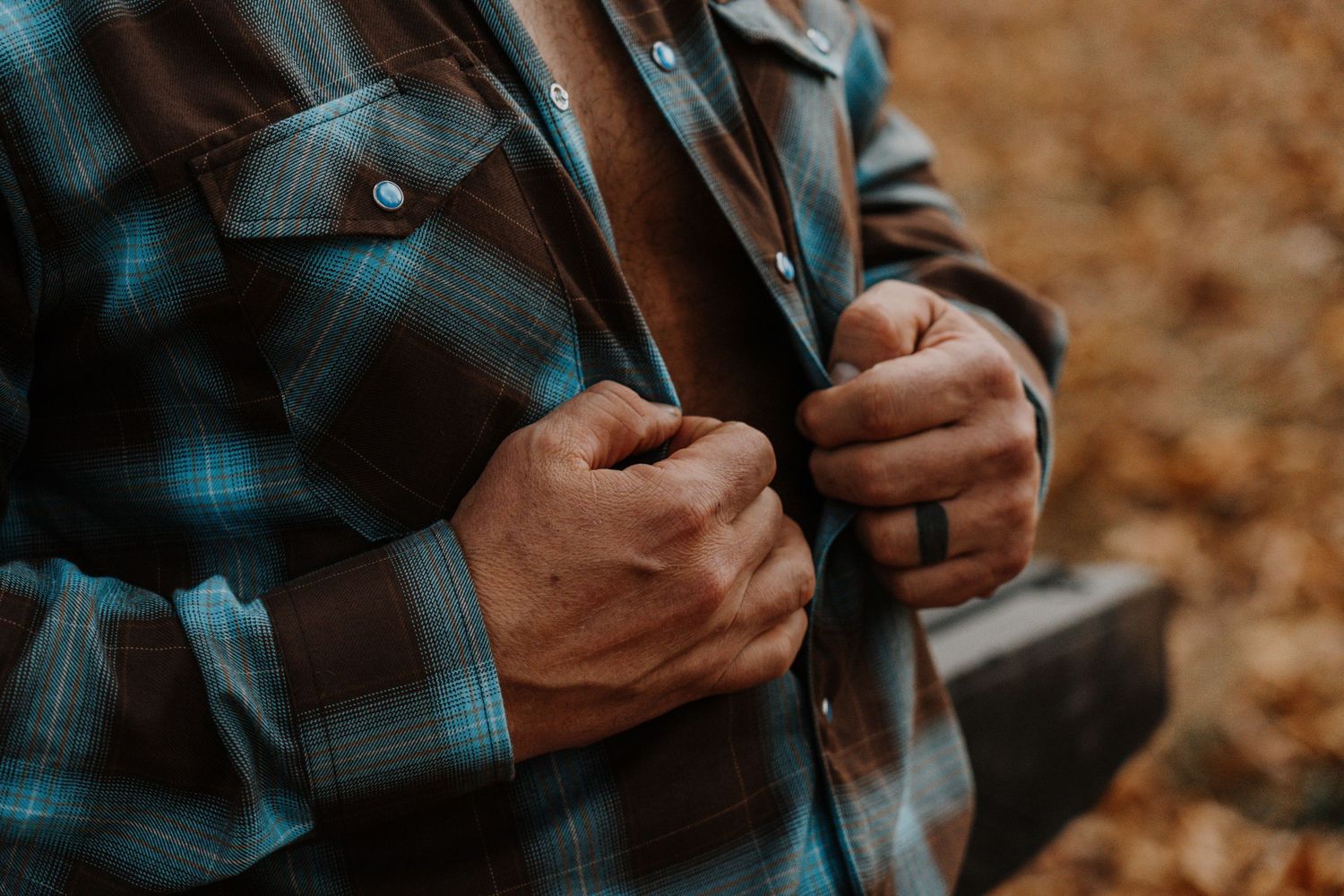 Close-up view of hands buttoning up a teal and black plaid flannel shirt against an autumn background.