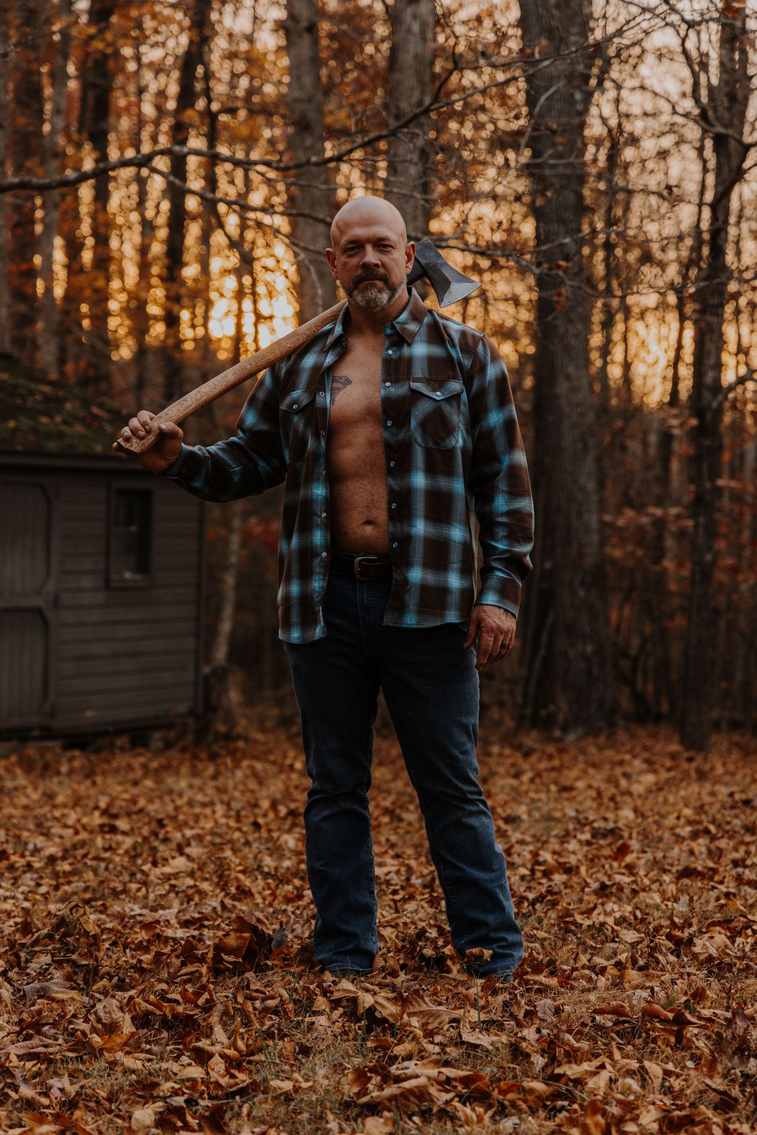 A person in an unbuttoned plaid shirt stands in autumn woods holding an axe at sunset with fallen leaves on the ground.