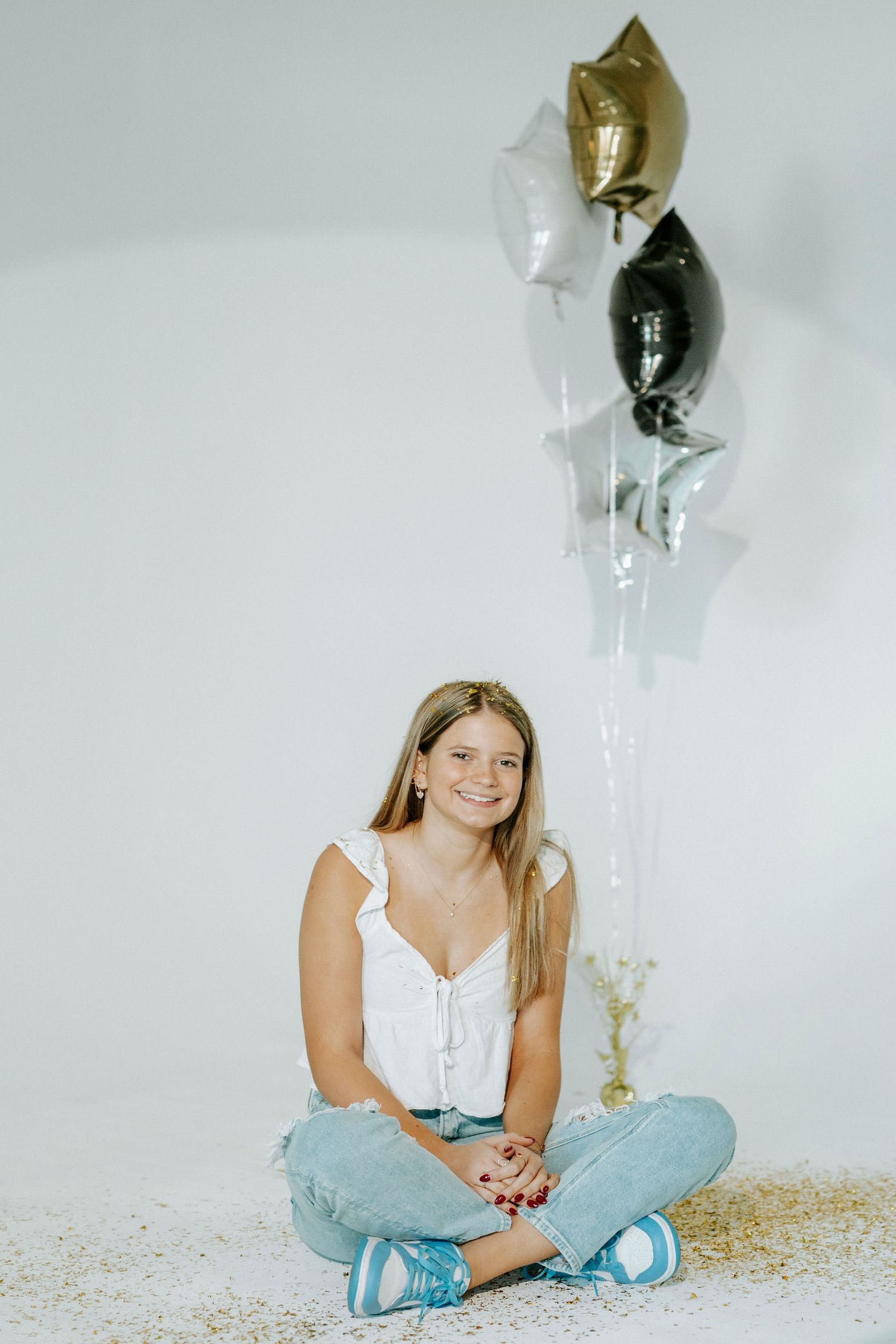A person in a white top and light blue pants sits on the floor holding shiny metallic balloons against a white background.