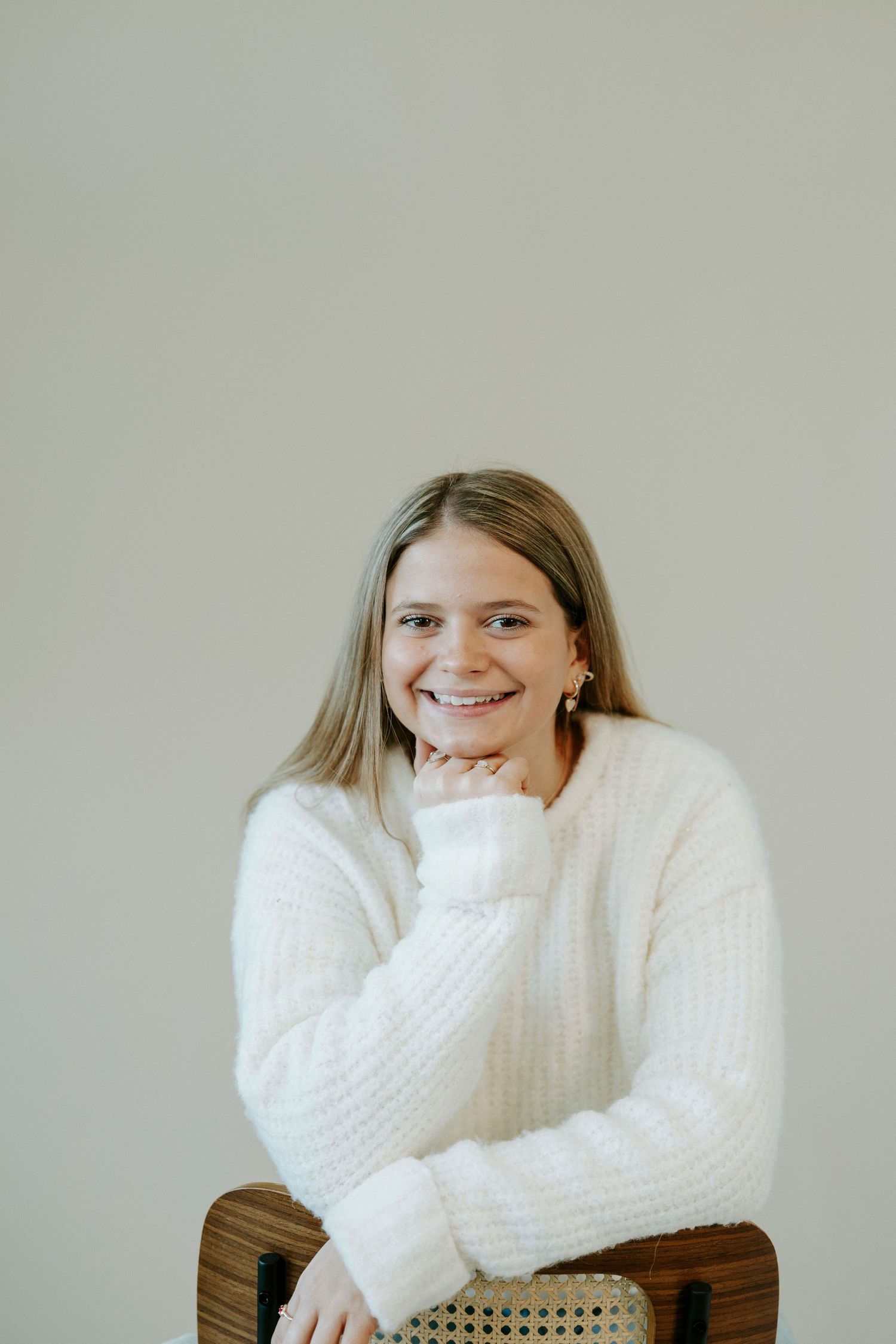 Someone wearing a cozy white knit sweater sits in a wooden chair against a neutral wall.