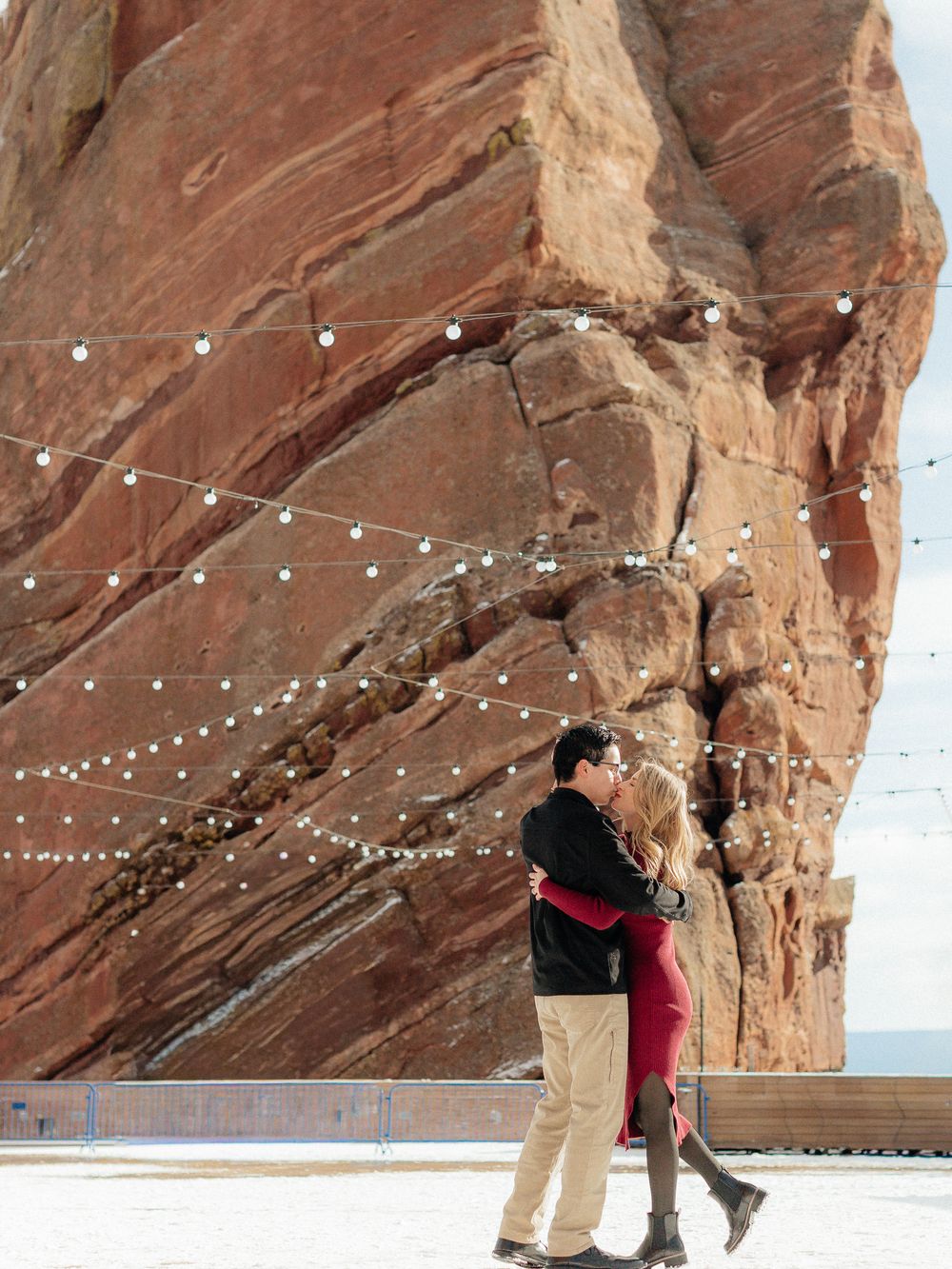 A couple shares a romantic moment under string lights against a dramatic red rock canyon wall at sunset.