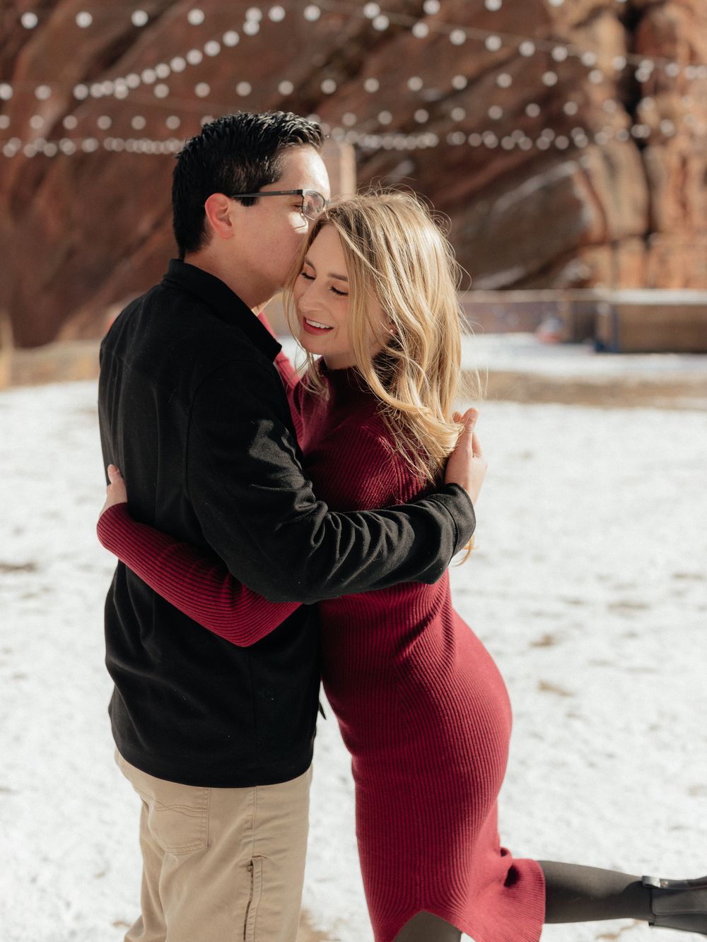 A couple embraces in a romantic winter scene with twinkling lights and snow in the background.