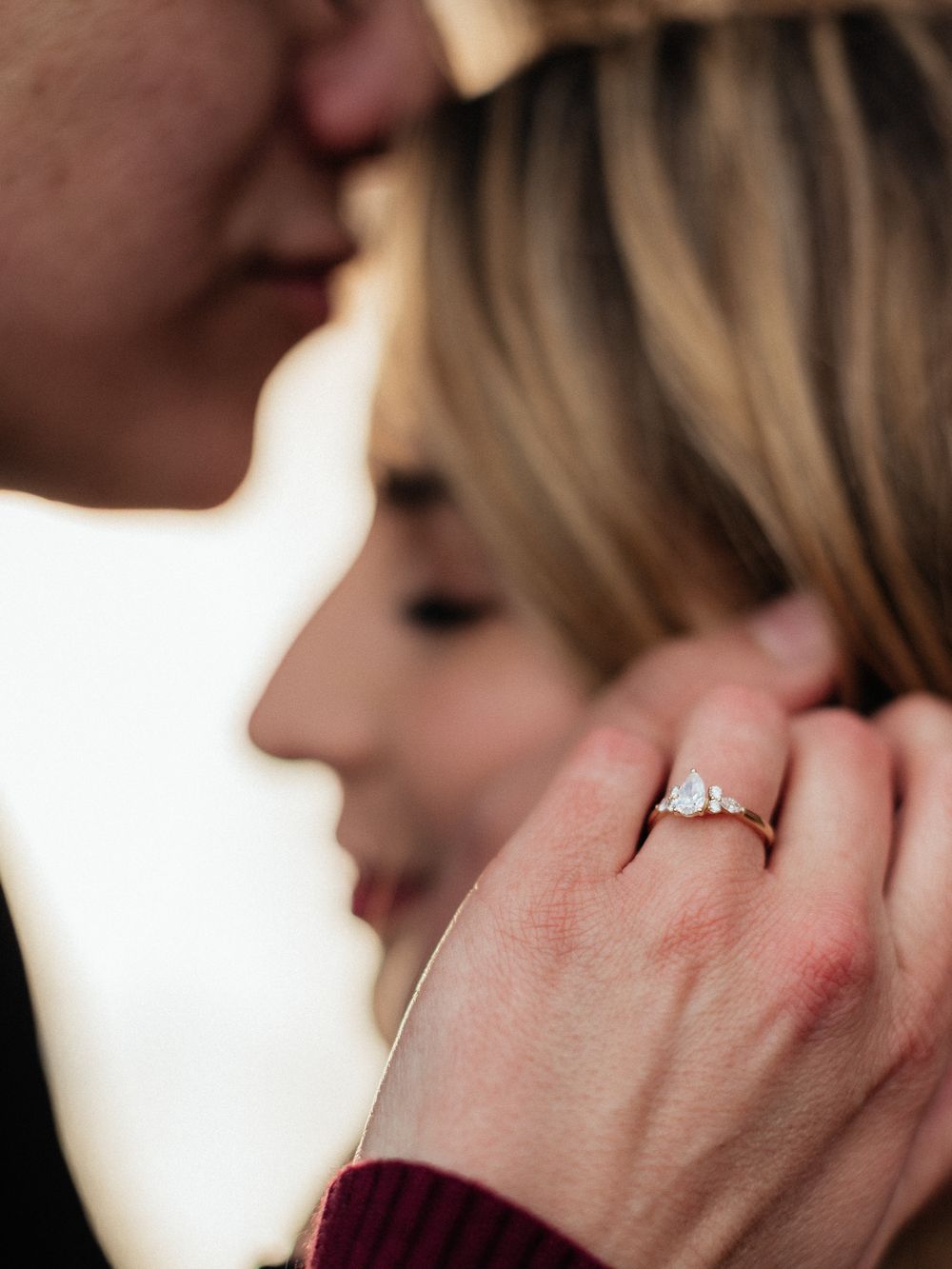 Close-up photo showing an engagement ring on a hand while a couple shares an intimate moment.