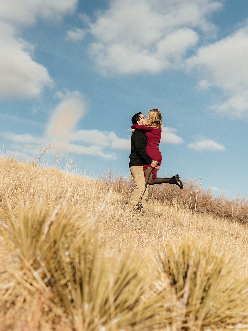 A joyful couple embracing in a golden wheat field under a bright blue sky with fluffy white clouds.