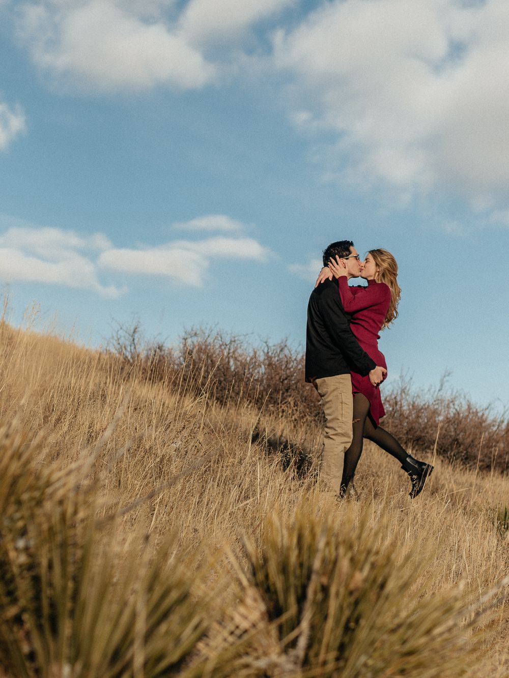 A couple shares an intimate moment together while standing in tall golden grass on a hillside under blue skies.