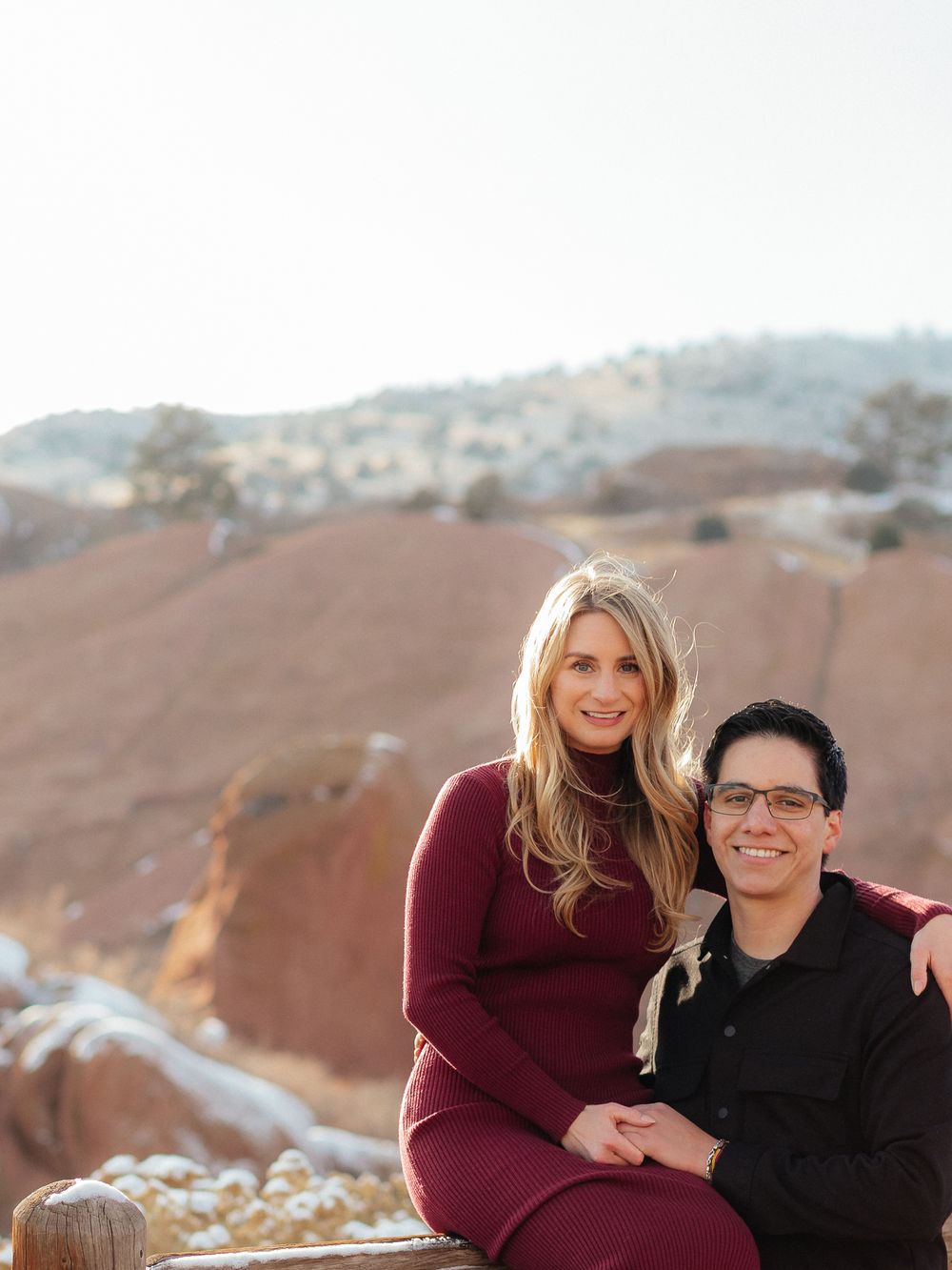 A couple sits together on a rocky overlook with desert mountains and canyons in the background.