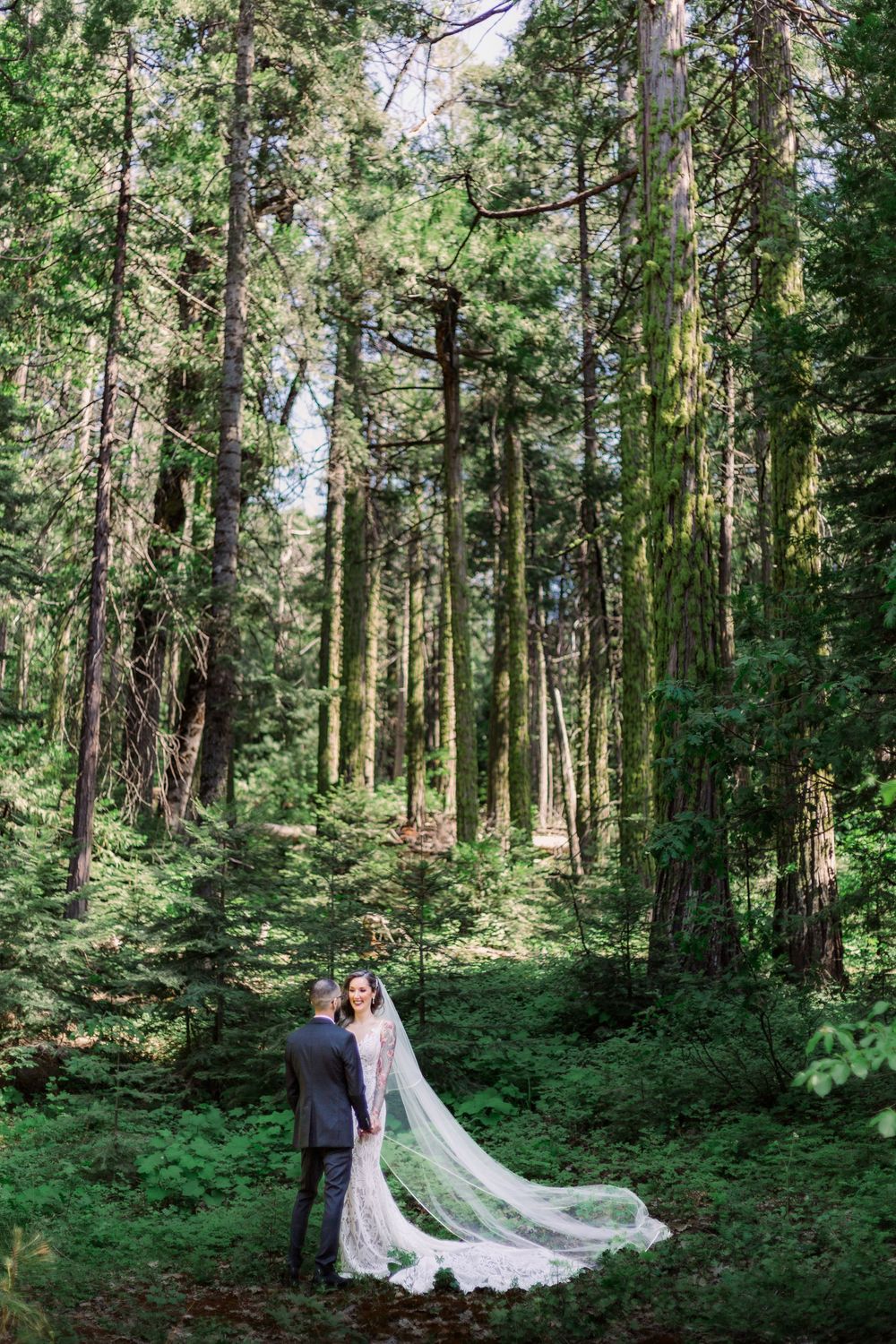 Newlyweds share intimate moment in enchanted forest setting with dramatic cathedral veil.