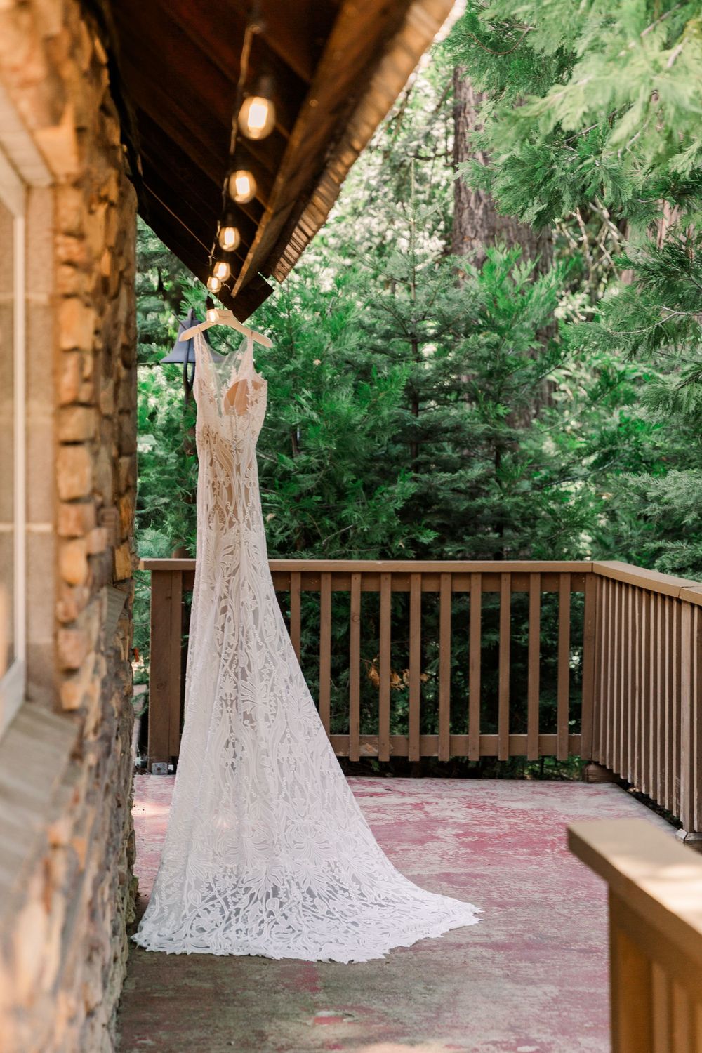 White lace wedding gown displayed on covered wooden porch with forest backdrop.