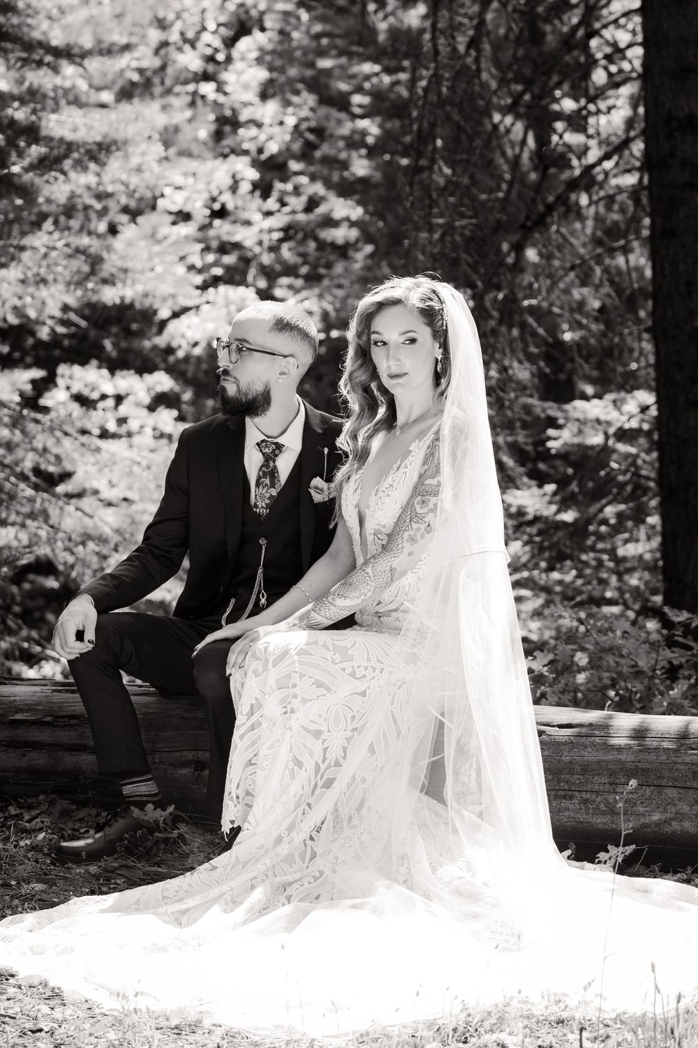 A black and white wedding photo of a couple sitting on a wooden bench in a forest setting.