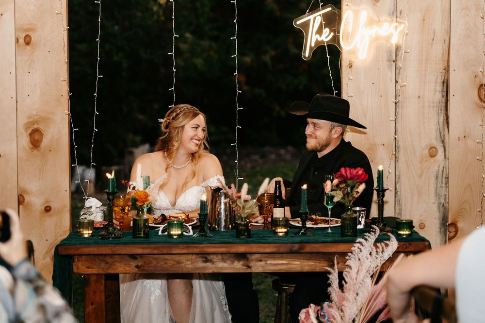 A rustic barn wedding reception with wooden walls, string lights, and guests gathered at long tables for dinner.