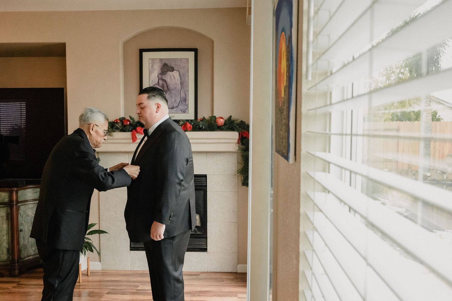Two people adjusting formal attire near a window in preparation for a wedding.