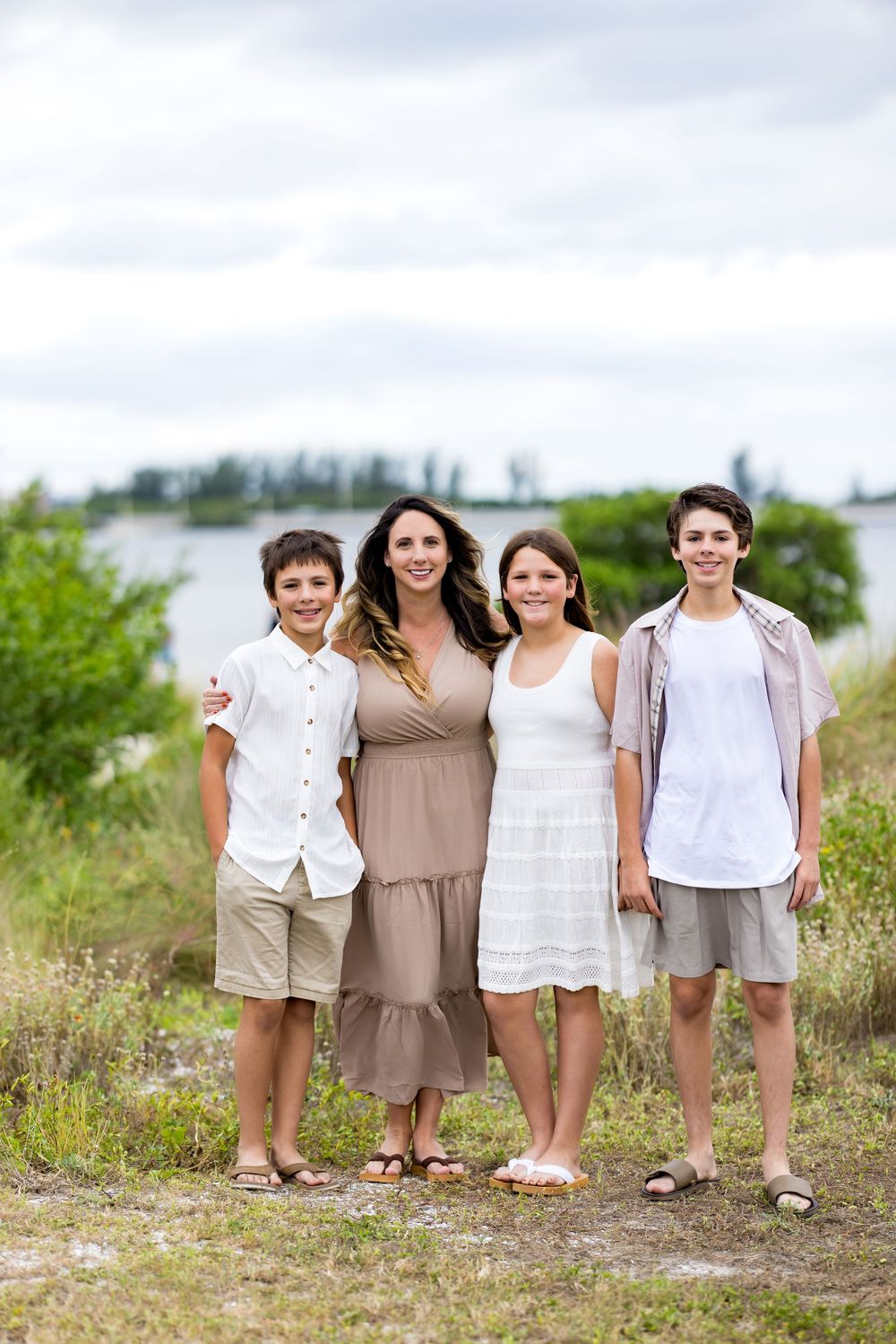 A family of four stands together outdoors on a grassy path near the water on a summer day.