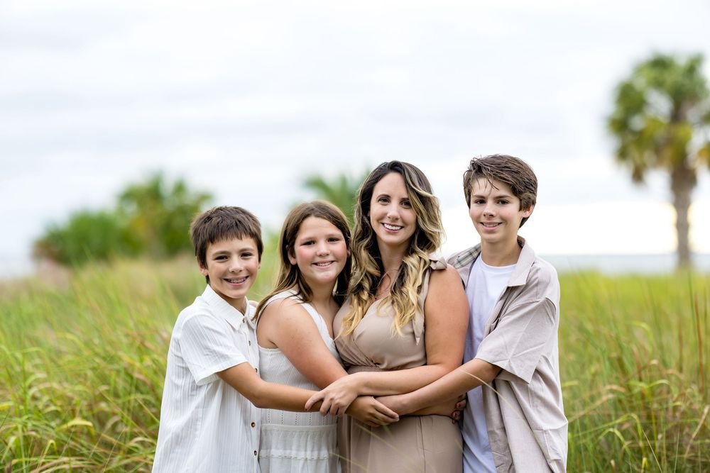 A family dressed in white and khaki outfits poses together on a grassy beach path during sunset.