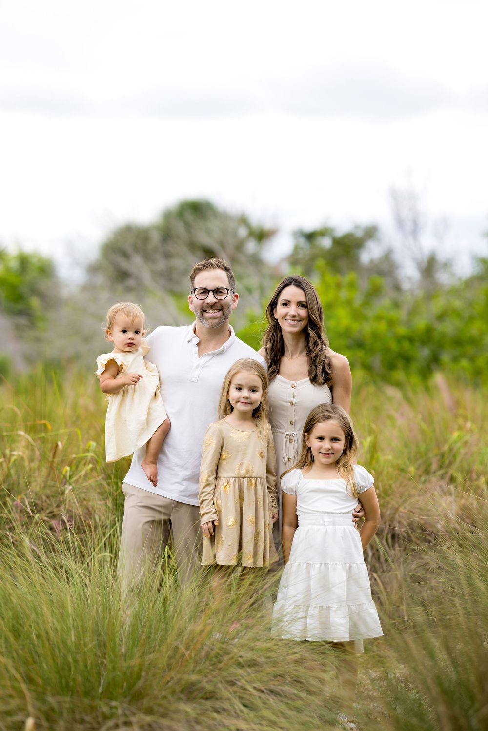 A family wearing coordinated white and beige outfits poses together in tall grass during an outdoor photo session.