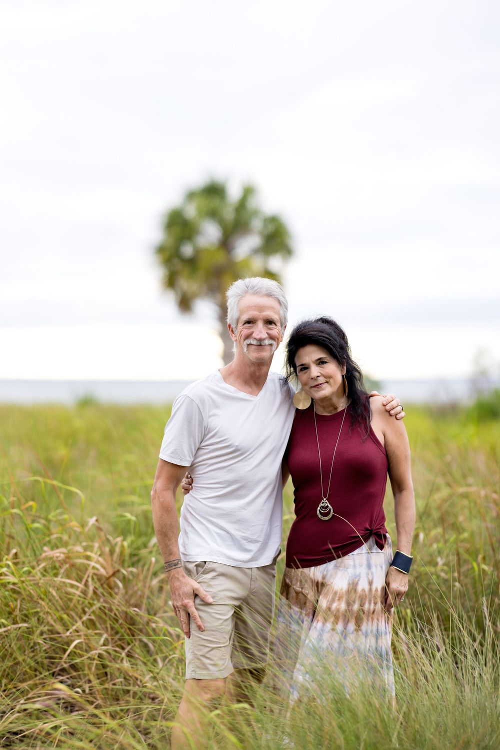 A couple poses together in a grassy field on a sunny day wearing burgundy and white casual attire.