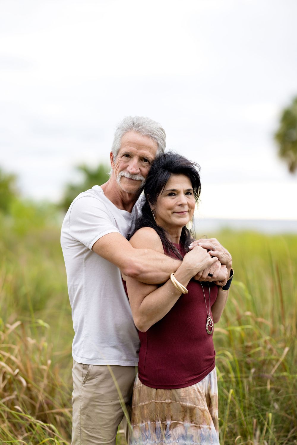 A couple embraces and laughs together in a sunny meadow with tall grass and greenery in the background.