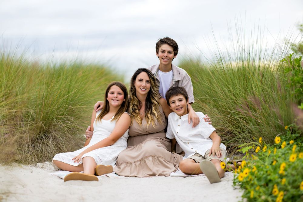 A family relaxes together on a sandy beach with tall grass and yellow wildflowers in the background.