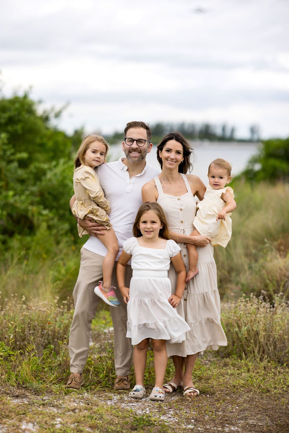A family group poses together outdoors in coordinating white and neutral outfits among wildflowers and natural scenery.