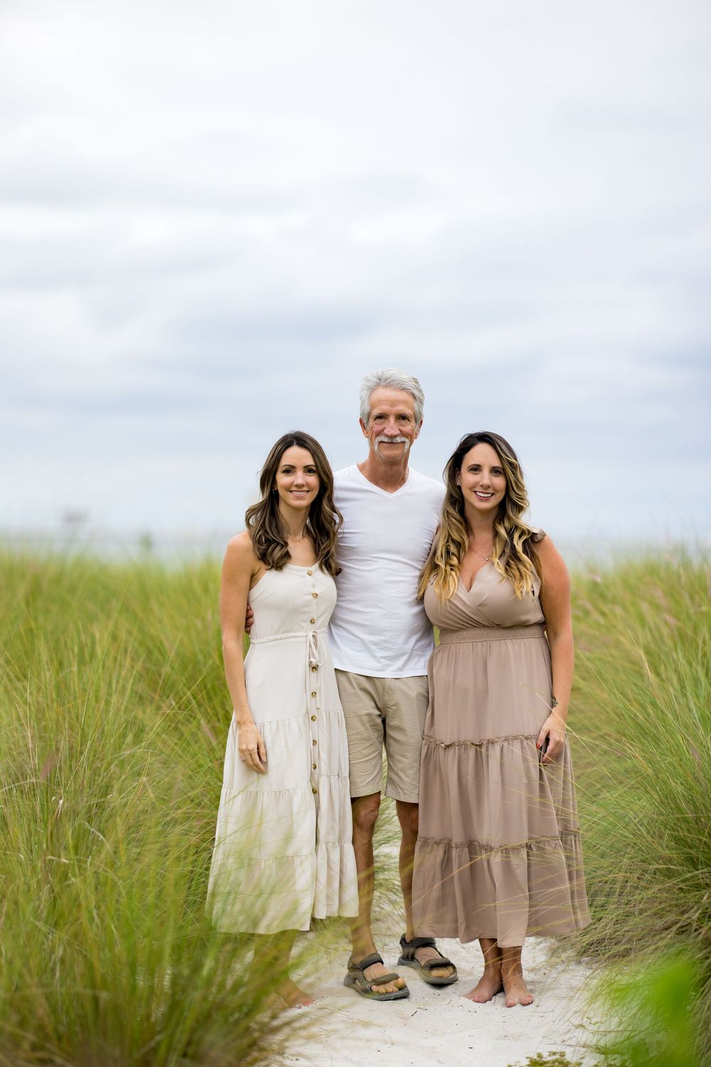 Three people stand together smiling on a beach path surrounded by tall grass and dunes on a cloudy day.