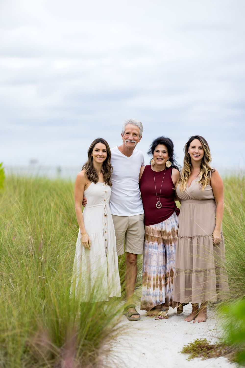 A group poses together on a sandy beach path surrounded by tall grass on a cloudy summer day.