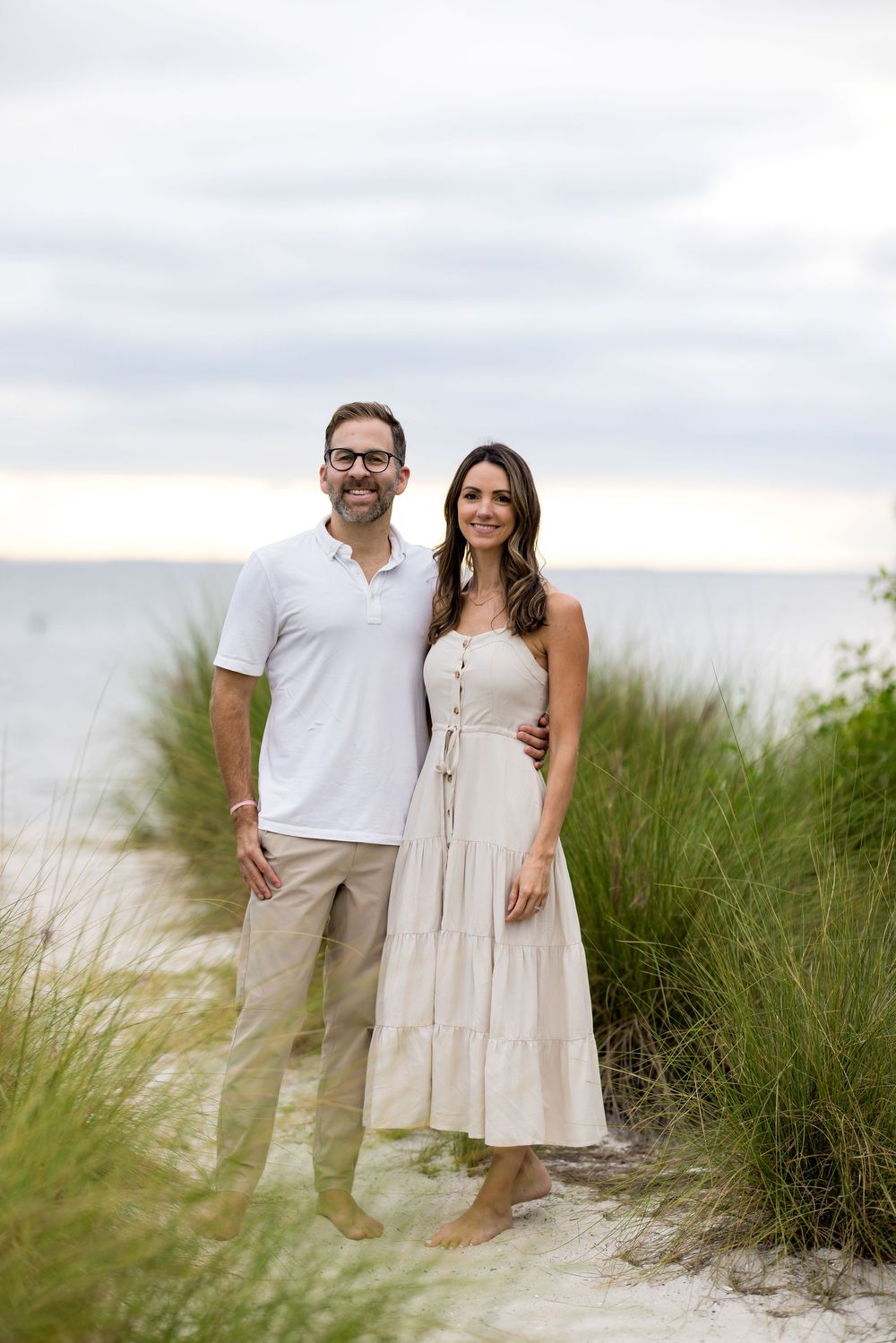 A couple in light-colored outfits stands together on a sandy beach path surrounded by tall beach grass.