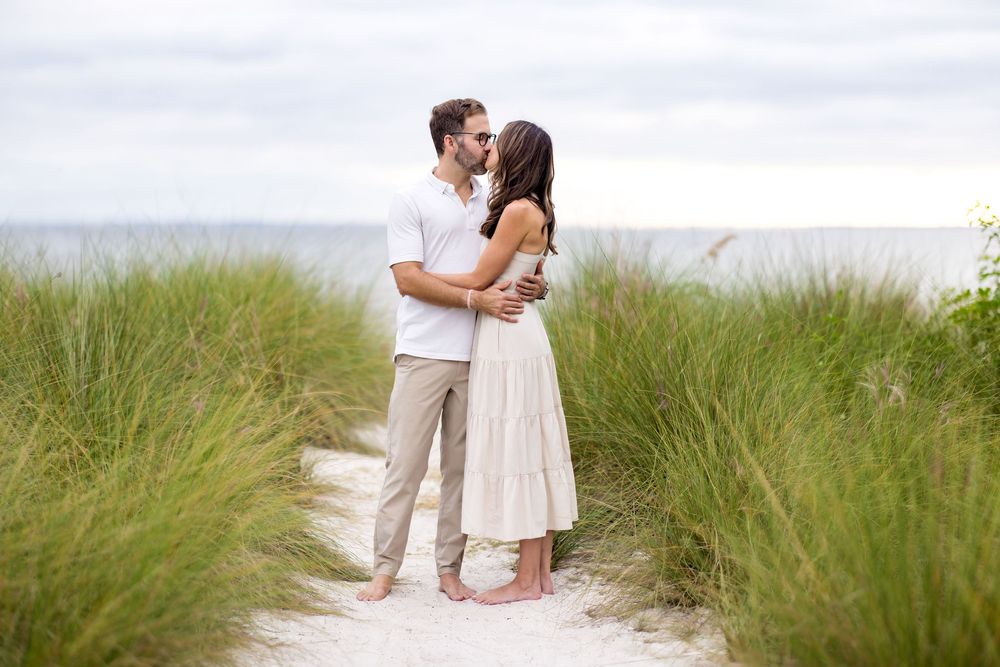 Romantic couple embraces on sandy beach path surrounded by tall seagrass during sunset.