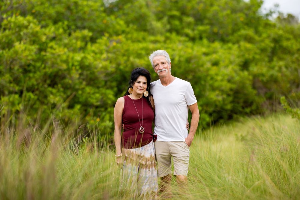 A couple stands together in a field of tall grass with green foliage in the background.