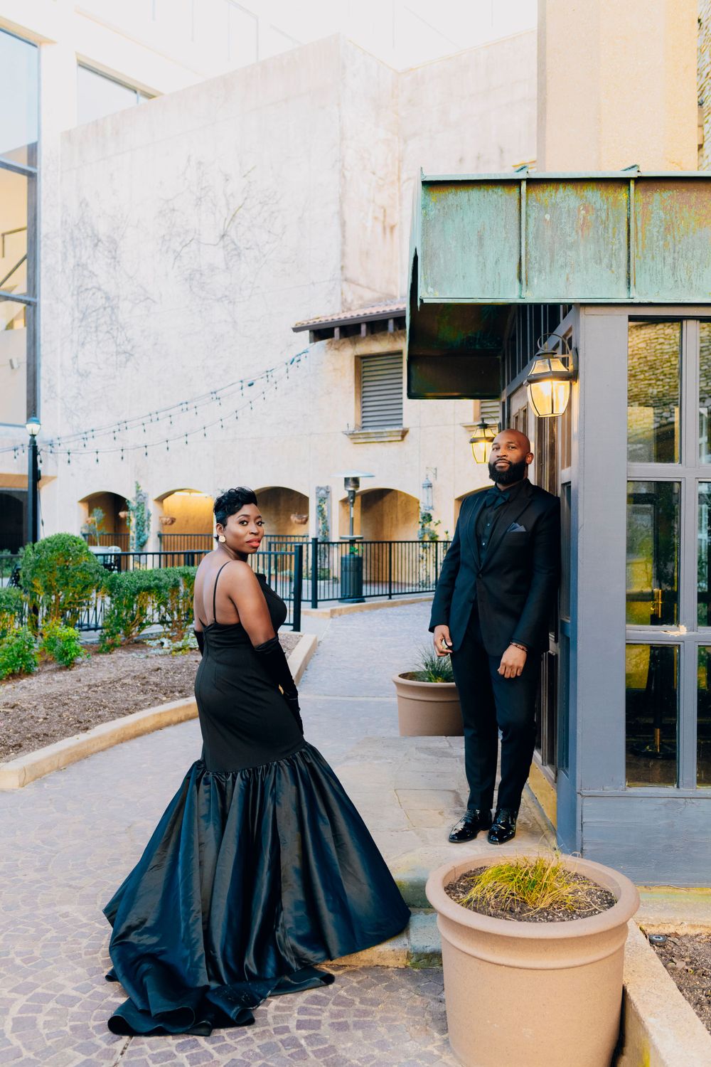 Couple in elegant black formal attire standing outside a Mediterranean-style venue with string lights overhead.
