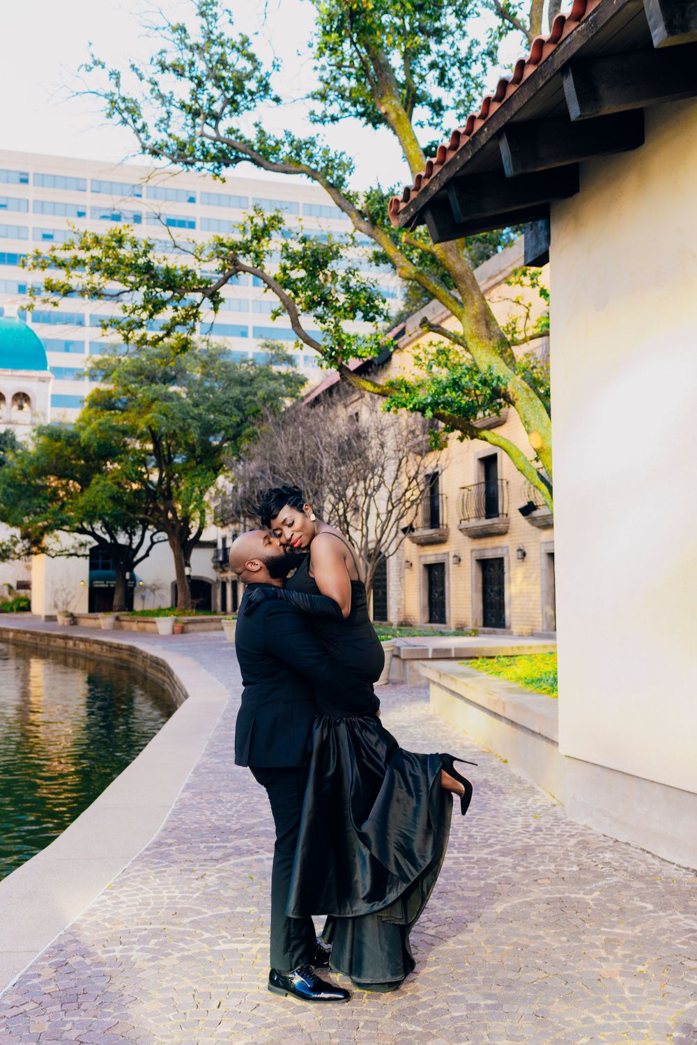 A couple in formal attire embraces by a reflective pool outside a Spanish-style building on a sunny afternoon.