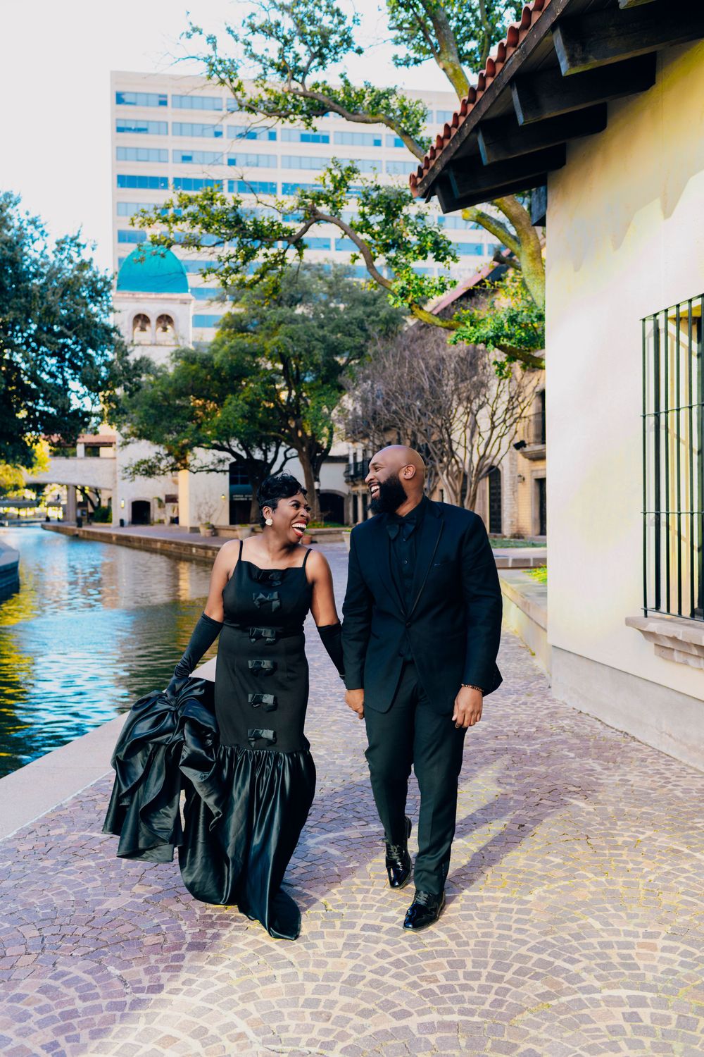 A couple in elegant black attire walks along a waterway with a modern building in the background.