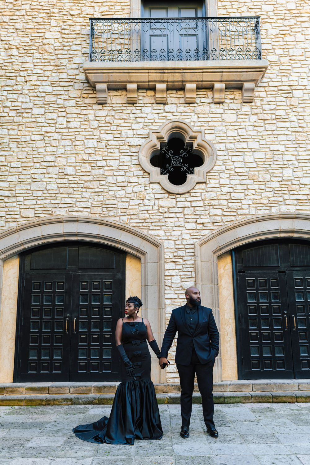 Elegant couple in black formal attire pose together against ornate stone wall with decorative doors and balcony.