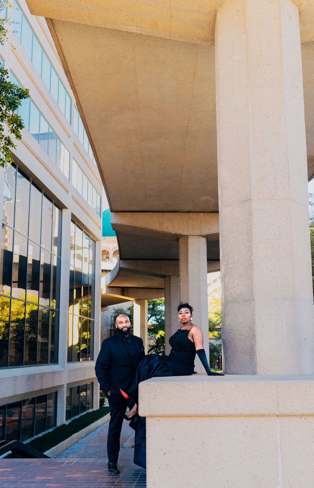 Two people stand in a modern architectural space with concrete columns and glass windows on a sunny day.