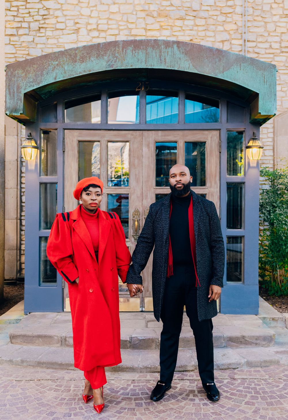 A couple stands in front of an arched doorway entrance, wearing coordinated red and black formal outfits.