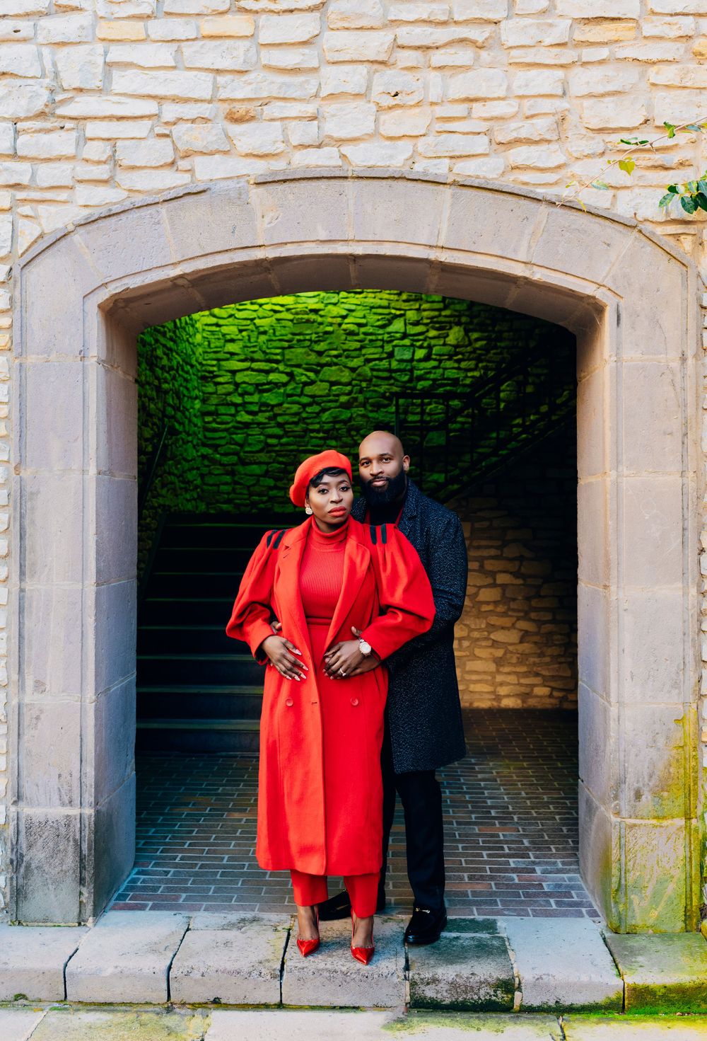 A couple stands in a stone archway entrance, with one wearing a striking red coat and beret while the other wears dark clothing.