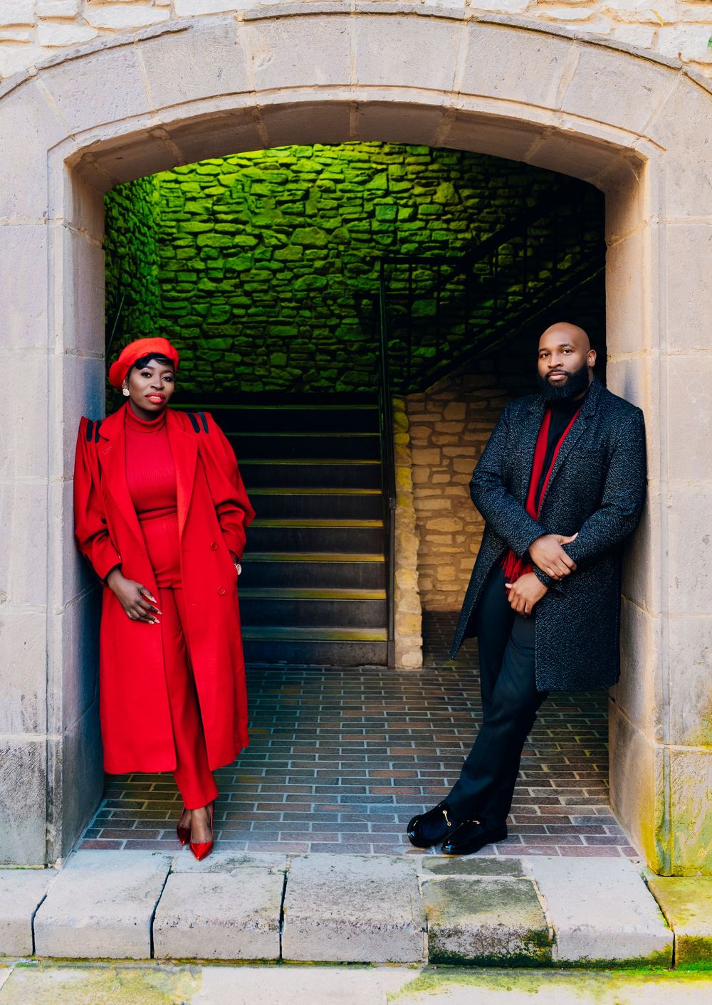 A stylishly dressed couple poses in a stone archway with green illuminated walls, contrasting red and black outfits.