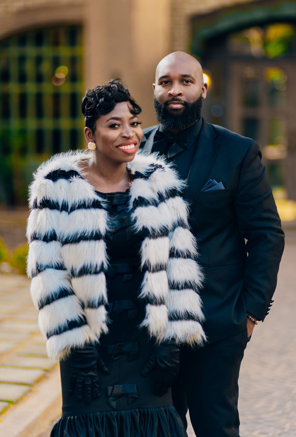 A couple poses outdoors wearing elegant winter attire including a striped fur coat and dark suit.